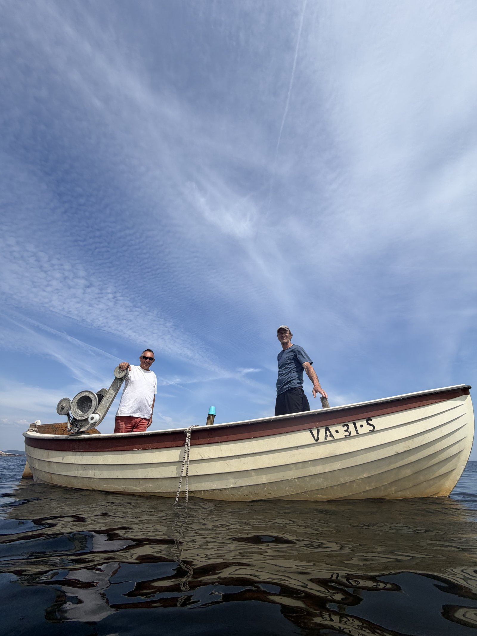 A wooden fishing boat with two guests aboard, reflected in calm summer water